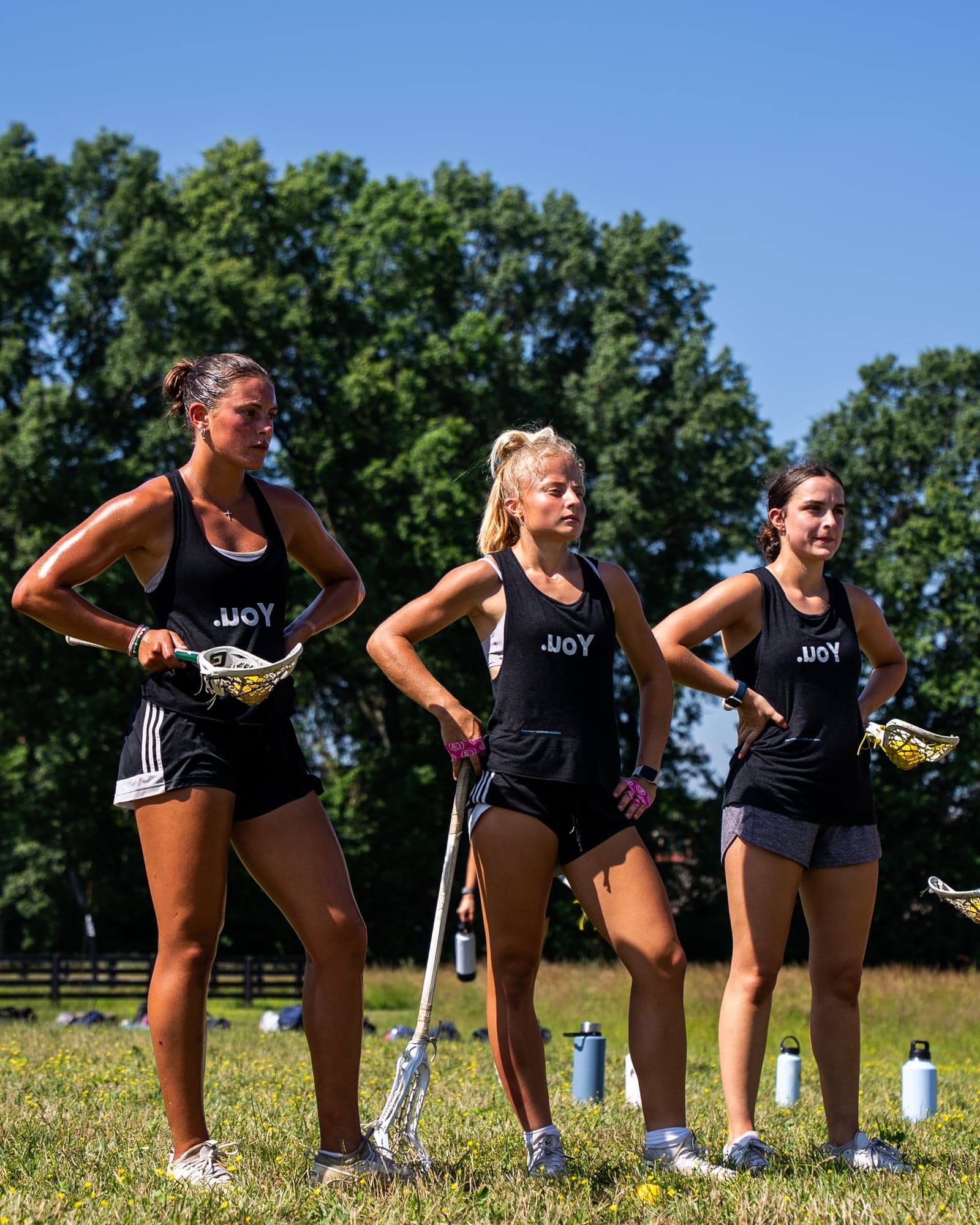 Three athletes in black .uoY tanks standing with lacrosse sticks on a sunny field